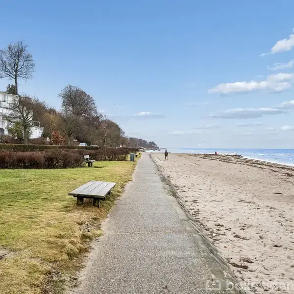 En gangsti løber langs en sandstrand ved havet. På den ene side er der græs og bænke, og på den anden side står huse og træer under en blå himmel.