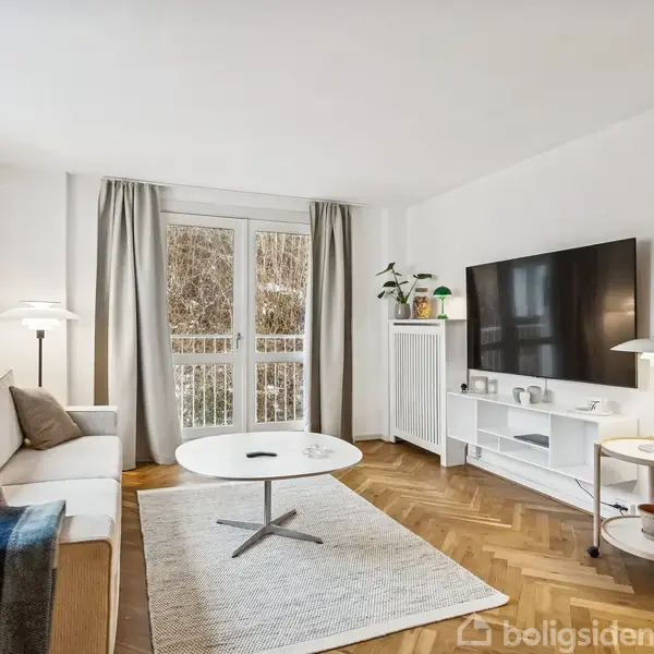 Stue med sofa og sofabord; en stor fladskærm foran hvid reol og ur på væggen; store vinduer med gardiner; gulvlampe og tæppe skaber hyggelig atmosfære.