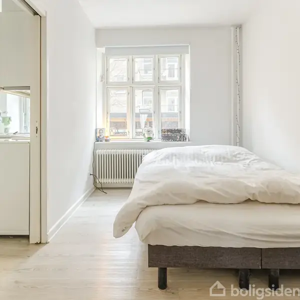 Seng står opredt i et lyst soveværelse. Det er placeret ved siden af et stort vindue, der giver naturligt lys. Rummet har trægulv og hvide vægge, med en radiator under vinduet.