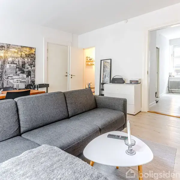 Grå sofa står i stue med sofabord, fjernbetjening og lysestage. Bagved ses et stort bybillede og spisebord med stole. Til højre er en åben dør mod et lyst rum.