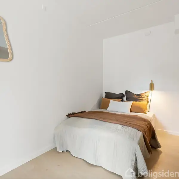 Et soveværelse har en opredt seng med hvide og brune sengetæpper. På væggen hænger et uregelmæssigt formet spejl. Rummet har lyse vægge og skabsplads.