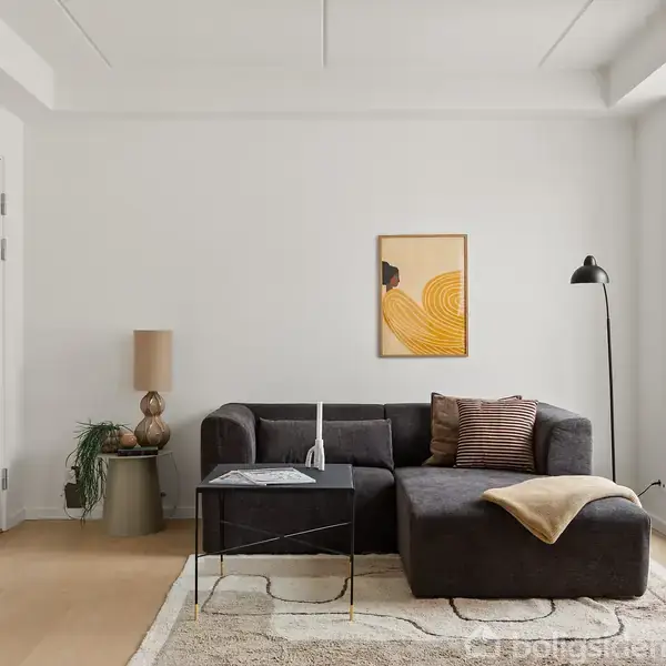 En sort sofa står i en lys stue ved siden af et lille bord med dekorative genstande. En gulvpude ligger på gulvet, og der er en plante i hjørnet ved vinduet. Væggen har et kunstværk med abstrakte former.