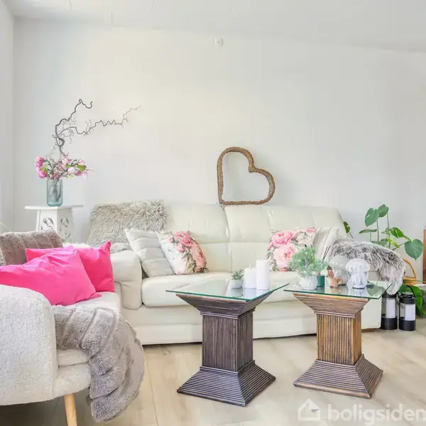 En stue med en hvid sofa dekoreret med blomstrede puder. To sofaborde med levende lys står foran sofaen. Til venstre er skønne planter og en sort reol ses i baggrunden.