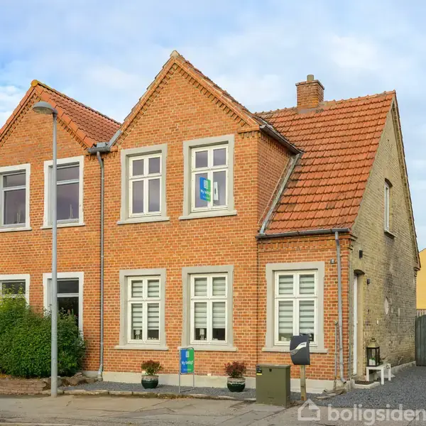 En rød murstensvilla med to etager står stille på en vej fyldt med vegetation. Huset har et tegltag og hvide vinduer, omgivet af buske og en asfaltsti.