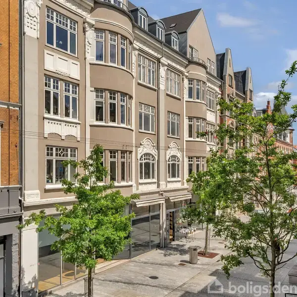 Etagebygning med klassisk arkitektur står langs en stille gade. Flere træer er plantet langs fortovet under en klar blå himmel.
