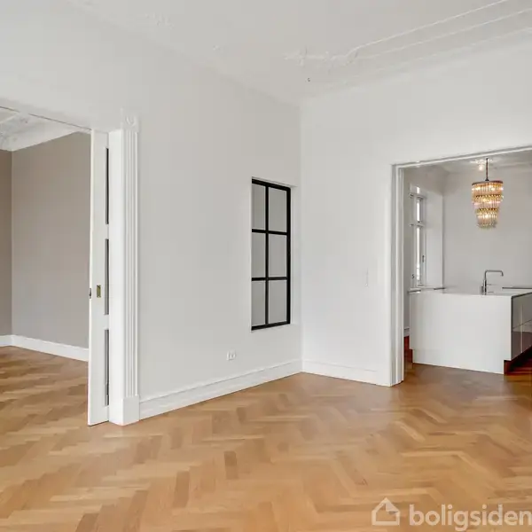 Et tomt rum med parketgulv åbner ind til to tilstødende værelser, heriblandt et køkken med moderne indretning. Rummene har høje, hvide vægge og et elegant, lyst design.