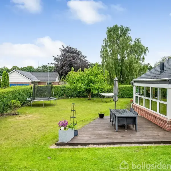 Hus med terrasse. En træplatform med bord, stole og parasol ved siden af et glashus. Trampolin og frodig have med græsplæne omgivet af hække og træer i baggrunden.