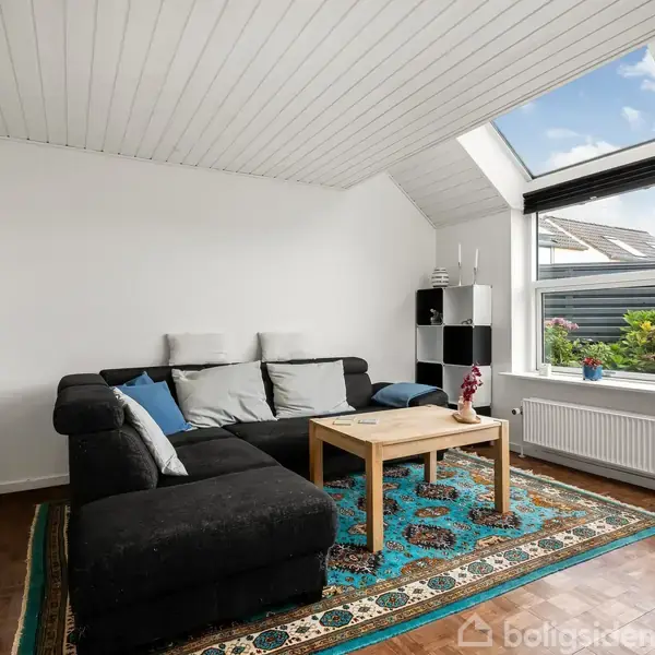 Sort sofa står i en stue, dekoreret med puder, foran et firkantet træbord. Store vinduer til højre giver udsigt til en have, mens hylder fylder venstre væg.