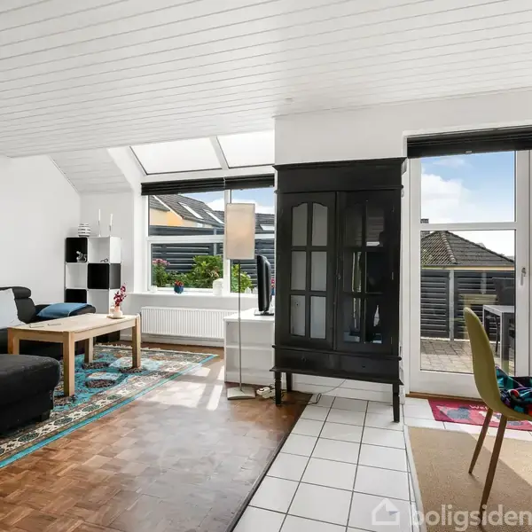 Sofa står i en lys stue med trægulv og tæppe, ved siden af et sort skab. Vinduer og fransk altan slipper dagslys ind, mens et lille bord og stole ses til højre.