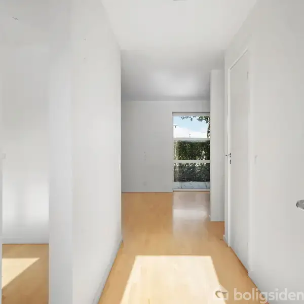 En lang gang med lyst trægulv fører mod en glasdør i det fjerne. Rummet er lyst med hvide vægge og døre på begge sider.