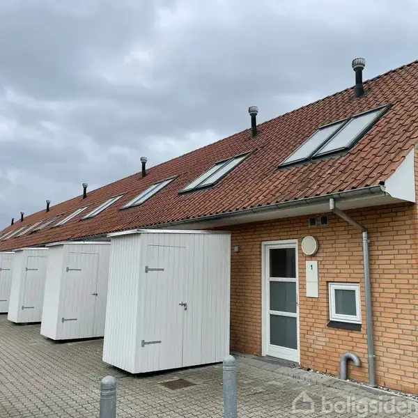 Rækkehus med rødt tag har flere døre og opbevaringsrum foran, placeret i et rækkehusområde. En skydedør bærer nummeret "1". Skyer dækker himlen.