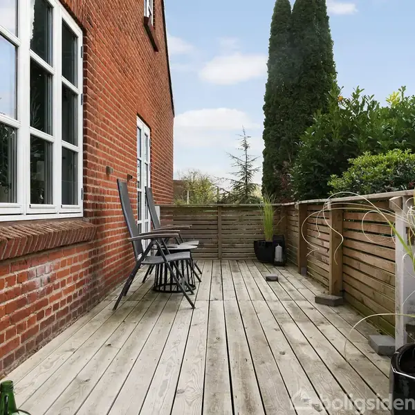 En terrasse med trædæk huser to sorte stole. Terrassen er op ad en rød murstensbygning og omkranset af træhegn. Planter ses i baggrunden ved siden af høje træer.