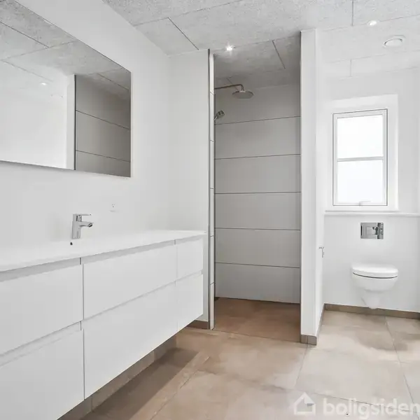 Badeværelse med hvidt skabsmodul og vask, spejl, bruseniche uden forheng og væghængt toilet. Lysegråt gulv og hvid væg i rummeligt miljø med stort vindue ved siden af toilettet.