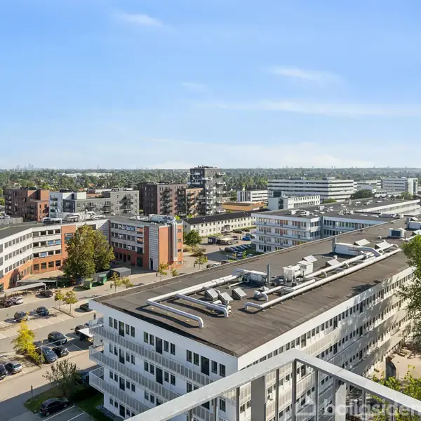 Bygninger i forgrunden strækker sig mod horisonten, omkranset af træer og veje med parkerede biler, under en klar blå himmel i et bymiljø.
