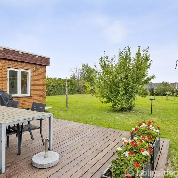 En terrasse med spisebord og stole står ved siden af et murstenshus. I haven er der en græsplæne med buske og træer. En flagstang står i baggrunden.