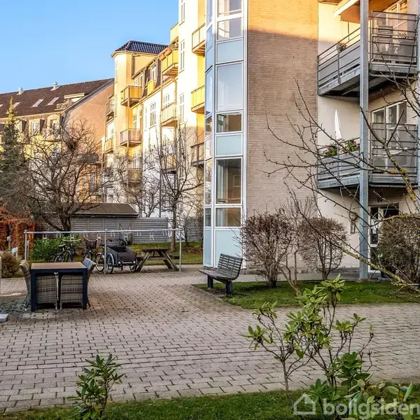 En lejlighedsbygning står stille i et solbeskinnet gårdområde med træer, bænke og cykler. Bygningen har altaner, og solen oplyser facaden bag en blå himmel.