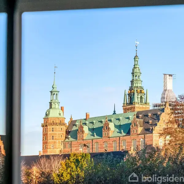 Et gammelt slot med tårne og et grønt tag ses udefra gennem en vinduesramme. I baggrunden er der en blå himmel og trætoppe.