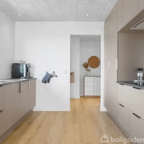Et moderne køkken med beige skabe langs begge sider, en kaffemaskine på bordet, og et vindue til venstre. Foran er en åbning til en anden lysfyldt gang med runde spejle.