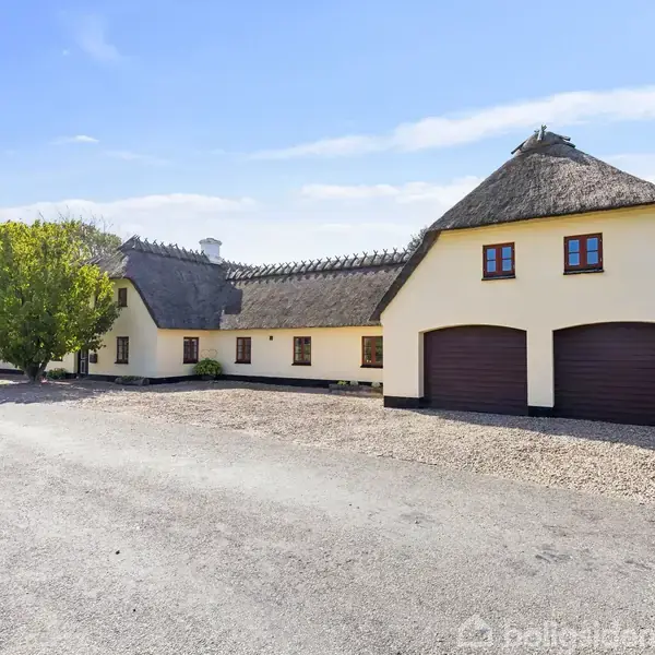 Lyst-gulmalet bindingsværkshus med stråtag, står stille i solrigt vejr ved grusbelagt indkørsel. To store garageporte ses til højre; træ markerer haven til venstre.