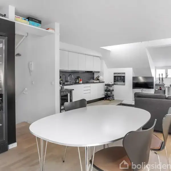 Et rundt bord med stole står i et moderne køkken-alrum. Køkkenet har hvide skabe og hvidevarer, og en grå sofa står nær et tv i baggrunden. Lysindfald fra ovenvindue.