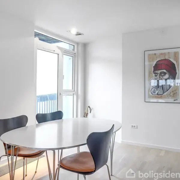 Ovalt hvidt bord med fire stole står i et lyst rum med et stort vindue. På væggen hænger et portræt af en mand med rød hue.