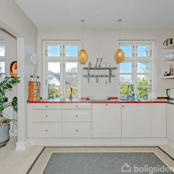Køkken med hvide skabe og rød bordplade, to hængelamper over bordet, planter i hjørnet, åbne hylder med køkkenredskaber, og to store vinduer med udsigt til en have.