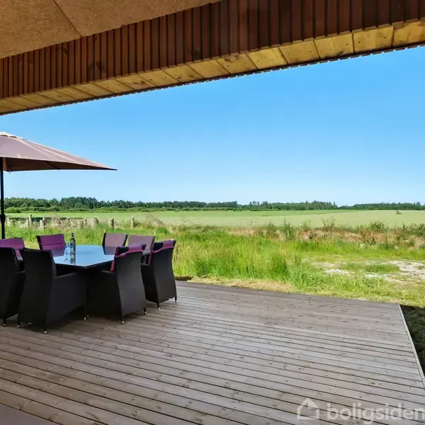 Terrassemøbler med parasol står på en træterrasse. I baggrunden ses en grøn mark under en klar blå himmel. Møblerne er arrangeret omkring et langt bord.