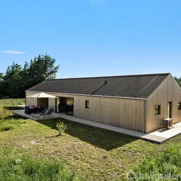 Træhus står stille på en stor, grøn grund med en terrasse til venstre. Omkringliggende natur består af græs og buske. Solrigt vejr med blå himmel uden skyer.