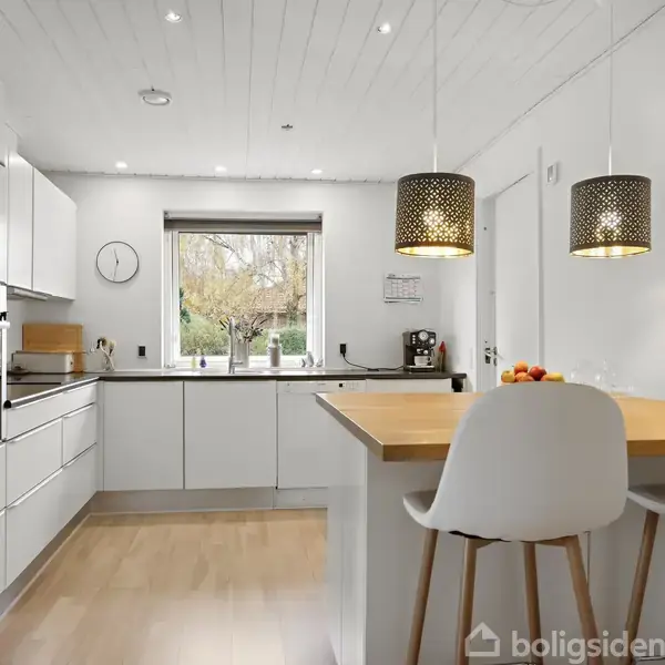 Hvidt, moderne køkken med hvide skabe og rustfrit stål apparater. To lamper hænger over en træbordplade med to hvide stole. Et vindue viser en have udenfor. En vægur hænger på væggen.