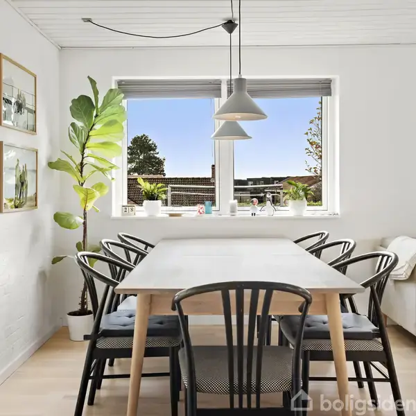 Et spisebord med sorte stole står i en lys stue. Planter pryder vindueskarmen, og en beige sofa står til højre. To lamper hænger over bordet.