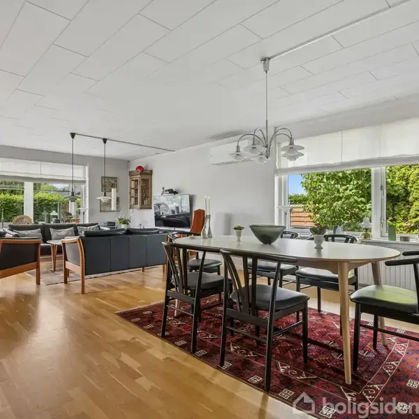 Spisestue med ovalt bord og sorte stole står på et rødt tæppe. Store vinduer viser en grøn have udenfor. Åbent køkken bagerst med moderne indretning og pendellamper.