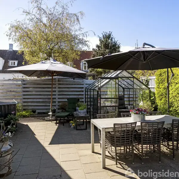 Terrasse med flisebelægning, hvor et havebord med stole står under en parasol. Et lille drivhus og planter ses i baggrunden. Sollys skaber skygger på terrassen.