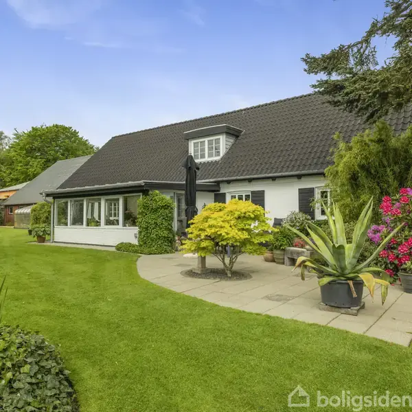 Et hvidt hus med sort tag står i en velplejet have. Grønne planter og farverige blomster omgiver terrasseområdet, hvor en parasol er opstillet.