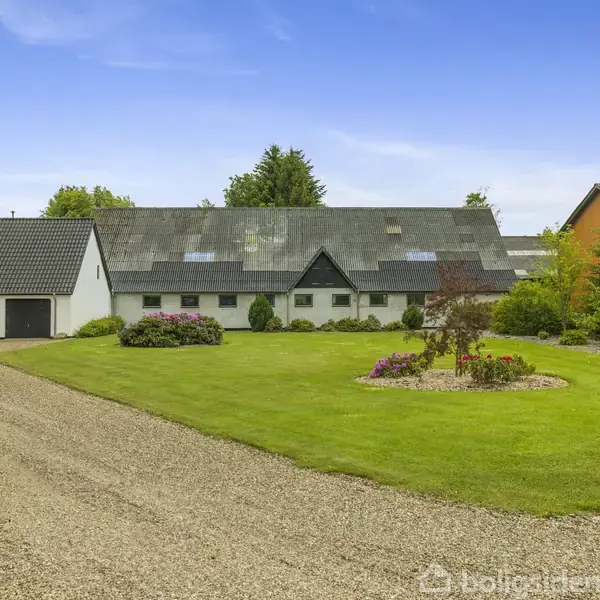 En hvid gårdbygning med sort tag står i en frodig have, omgivet af velplejet græs og blomsterbede. Til venstre ses en grusvej, der fører til en garage.