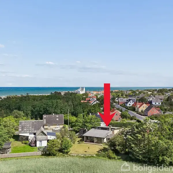 En bolig er markeret med en rød pil, omgivet af grønne træer og flere huse i et kystnært område. I baggrunden ses en kirke og havet under en let skyet himmel.