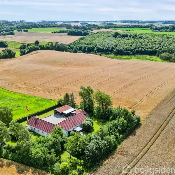 En hvid bygning med rødt tag ligger omgivet af træer midt i et stort landbrugsområde med marker. Flade, grønne landskaber og skove ses i baggrunden under en klar himmel.