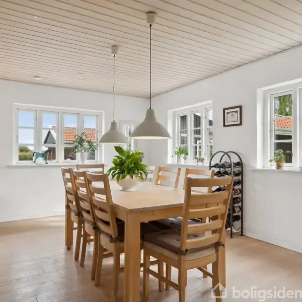 Et spisebord i lyst træ er omgivet af otte stole, placeret i et lyst køkken med trægulv. To lamper hænger over bordet, og tre vinduer giver udsigt til en have. En plante står centralt på bordet.