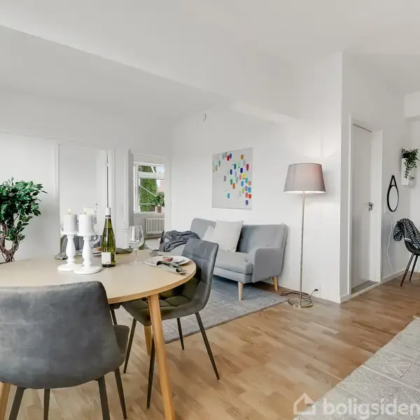 Rundt spisebord med stole står i lys stue. Lysestager og en flaske vin er på bordet. Sofa og gulvlampe ses ved væggen. Lys trægulv overalt.