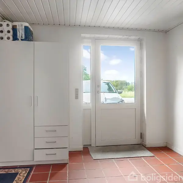 Et hvidt skab står i et rum med rødbrune fliser, tæt på en glasdør der fører ud til en grøn have. Toiletpapir er ovenpå skabet.