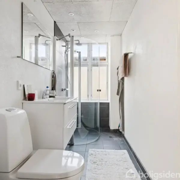 Toilet placeret i et lyst badeværelse ved siden af et skab med håndvask og spejl. Brusekabine med glasdør i baggrunden. Flere hylder og kroge langs væggen.