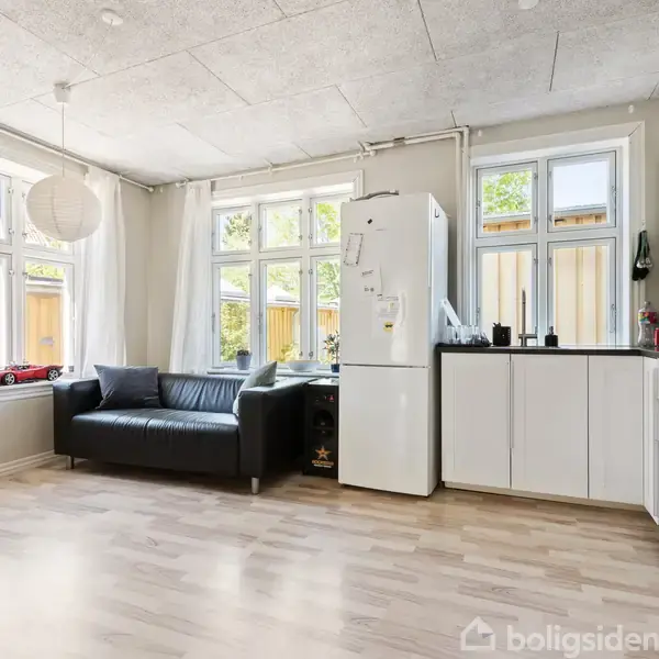 Sort lædersofa står stille ved to vinduer, der giver lys til køkkenet udstyret med hvide skabe og køleskab. Trægulv og lys pendellampe skaber en rolig atmosfære.