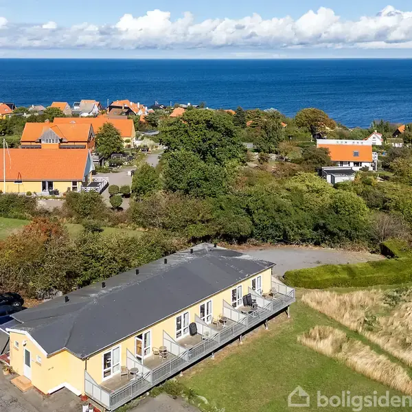 Bygning med gule vægge og sorte tage står stille blandt grønne buske og træer, omgivet af andre huse med orange tage. Havet ses i baggrunden under en skyet himmel.