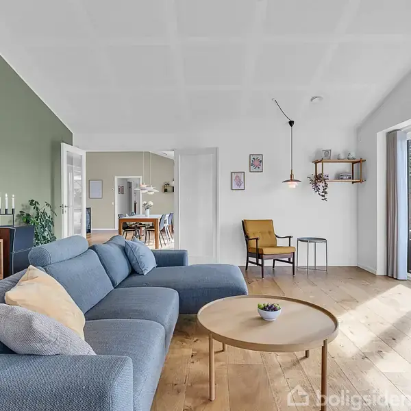 Blå sofa står i stue med trægulv, flankeret af et rundt sofabord. Til højre indgang til terrasse. Grøn væg med billeder og hylde, spisebord kan ses i baggrunden.