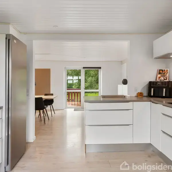 Et moderne køkken med hvide skabe og stålgrå køleskab. Køkkenet fører til en spisestue med sorte stole og et bord. Udsigt til grønne omgivelser gennem døren.