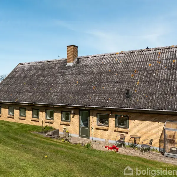 Et langstrakt, gult murstenshus med gråt, skrånende tag har en skorsten. Foran huset står et lille bord med to stole og en cykel. Græsplæne omgiver huset.
