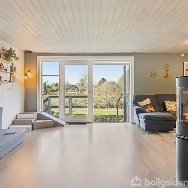 Stue med brændeovn til højre og stor hjørnesofa til venstre. Store vinduer åbner mod en terrasse med grøn udsigt. Hylder med planter på væggen og lyse trægulve.