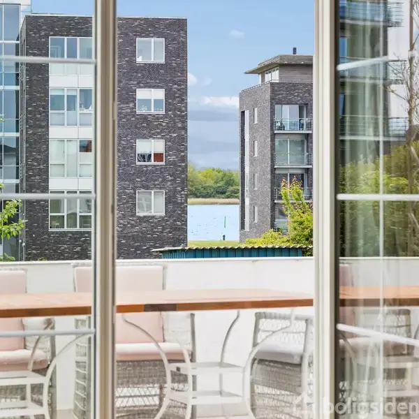 Glasdør viser udsigt til en altan med møbler og en lang bordplade. Baggrunden består af moderne lejlighedsbygninger og et vandområde, omgivet af træer under en klar himmel.
