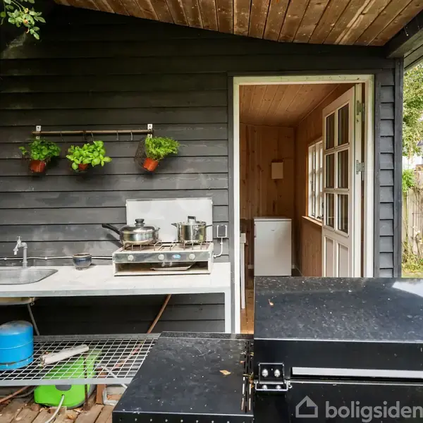 Udekøkken med sort væg, indeholder vask og kogegrej. Til venstre ses planter, til højre en åben dør til træsidedæk. Omkring er der et hegn og grønt græs.
