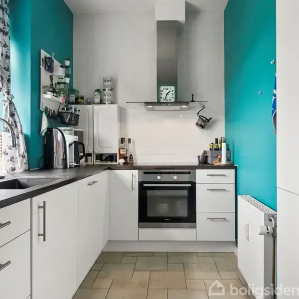 Et moderne køkken har hvide skabe og skuffer. Ovn og emhætte er centralt placeret. Grønne vægge og flisegulv omkranser rummet. Vindue til venstre med sort-hvide gardiner.