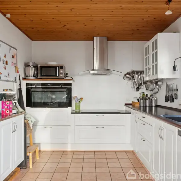 Et hvidt, moderne køkken med indbyggede borde og skabe. En ovn og mikrobølgeovn er integreret. Redskaber hænger på væggen. Trægulv og loft skaber en hyggelig atmosfære.